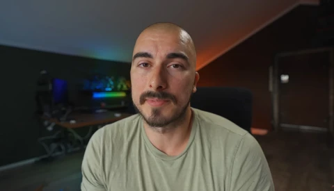 Man with shaved head and goatee wearing light gray v-neck t-shirt in dimly lit room.