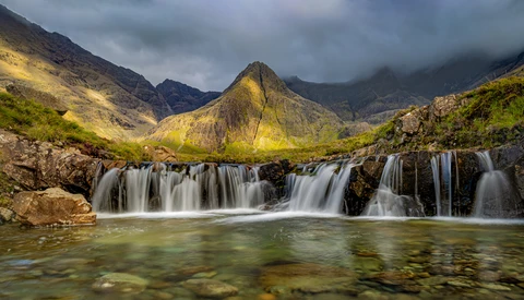 Cascading waterfall flows across layered rock formations into an emerald pool, with a distinctive peaked mountain rising beyond under dramatic cloudy skies.