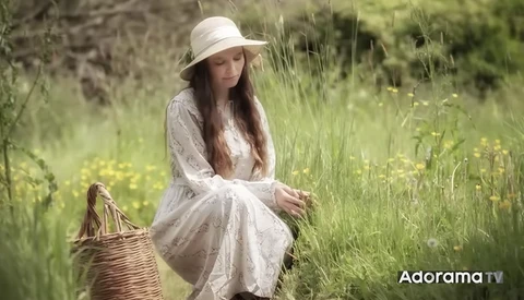 Woman in white dress and hat crouching in wildflower meadow beside wicker basket.