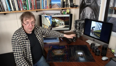 Photographer at desk reviewing images on monitor with camera equipment and shelving visible.