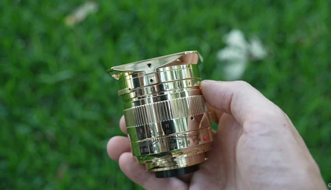 Hand holding a polished brass lens barrel with ribbed detailing and a wooden focus ring against blurred green foliage.