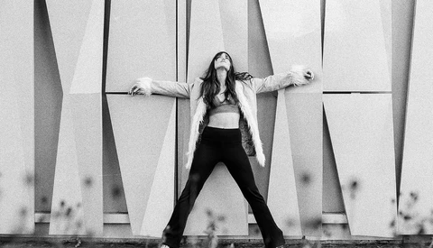 Woman in crop top and black pants striking a wide stance against geometric concrete wall panels.