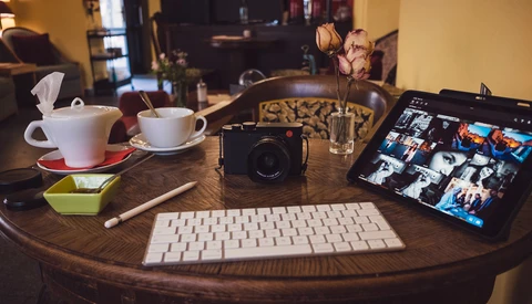 Photographer's desk setup with camera, keyboard, tablet displaying images, and coffee cups on wooden round table.