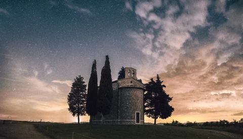 Cypress trees flanking a historic stone chapel at dusk beneath a starlit sky.