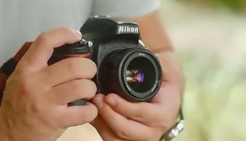 Hands holding a Nikon DSLR camera with a wide-angle lens against a blurred green background.