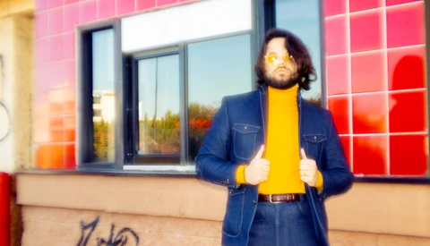 Person in denim jacket and yellow shirt standing against pink tiled wall with windows.