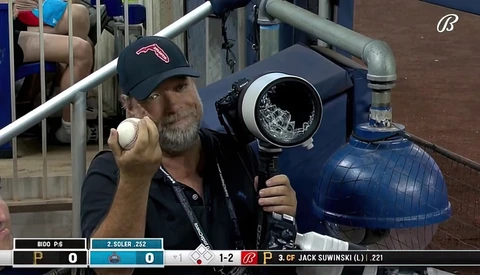 Man holding baseball and camera equipment at stadium during game broadcast.