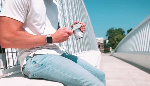 Person in white shirt holding smartphone while sitting outdoors against corrugated metal buildings.