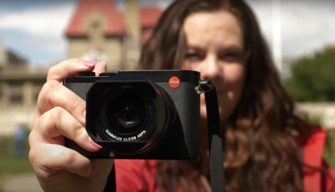 Woman holding a Leica camera toward the viewer, with a historic building blurred in the background.