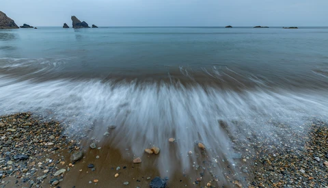 Long-exposure photograph of waves washing across a pebble beach at dusk, with rocky outcrops visible in the calm sea beyond.