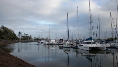Sailboats moored in calm harbor water with perfect mirror reflections under overcast sky.