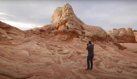 Photographer in dark clothing standing on layered sandstone formations, framing a tall rock pinnacle with camera.