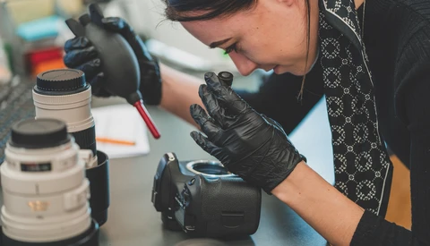 Photographer in black gloves examining a camera lens at a workbench with equipment and lenses nearby.