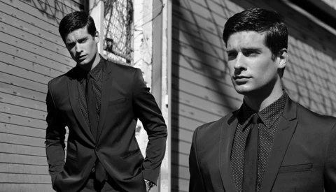 Two men in dark blazers and patterned shirts posed against an urban brick wall in black and white.