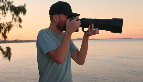 Male photographer holding a telephoto lens to his eye during golden hour near water.