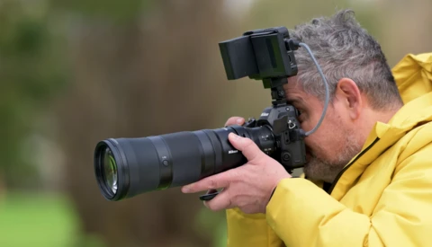 Photographer in yellow jacket holding camera with telephoto lens and electronic viewfinder.
