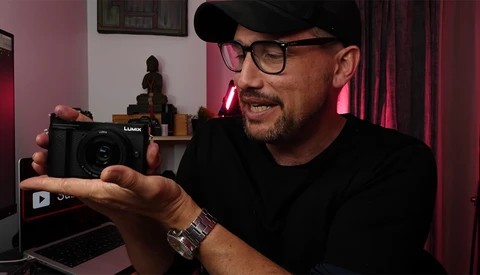 Photographer holding a Lumix compact camera while reviewing footage on a laptop in a studio setting.