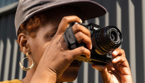 Photographer holding a rangefinder camera up to their face, wearing a cap and hoop earrings against a striped metal background.