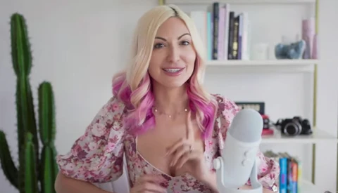 Woman with blonde and pink hair holding a white cylindrical microphone in a bright home studio setting.
