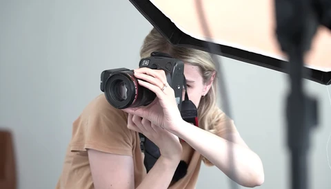 Photographer holding a mirrorless camera with telephoto lens to her eye while shooting in a studio setting.