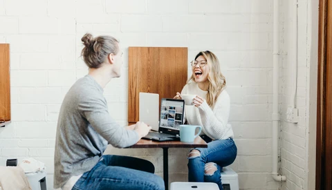 Two women sitting at a small table having a conversation and laughing in a bright studio space.