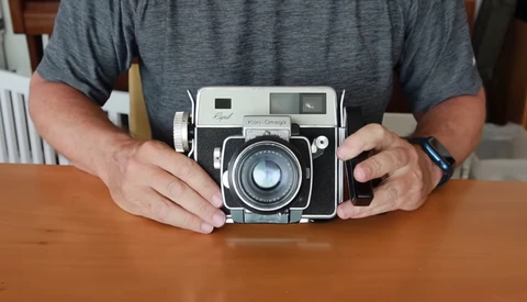 Person holding a vintage rangefinder camera with chrome and black body on wooden table.