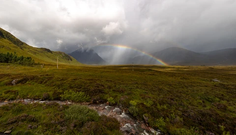 Double rainbow arcing over a vast green valley with mountains and dramatic storm clouds.