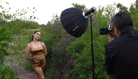 Photographer shooting a woman in bronze-toned clothing outdoors using a large parabolic softbox reflector.