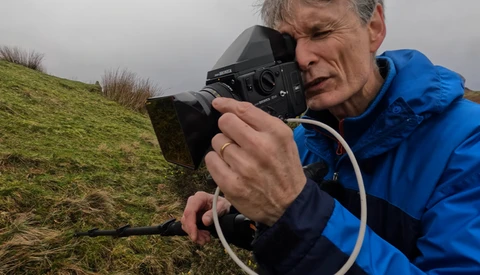 Photographer in blue jacket holding film camera to eye while composing shot on hillside.
