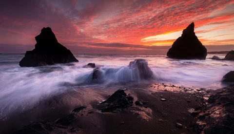 Dramatic coastal sunset with two dark rock formations silhouetted against an orange and purple sky, waves washing across dark sand in the foreground.