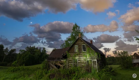 Abandoned wooden house surrounded by overgrown vegetation under a dramatic cloudy sky.