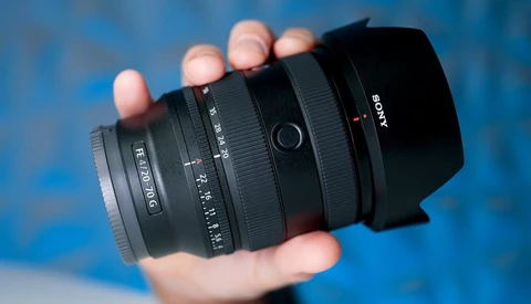 Hand holding a Sony camera lens with black barrel and hood against blue background.