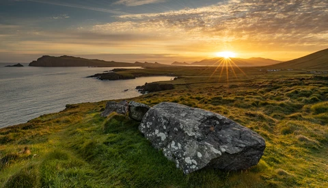 Lichen-covered boulder on grassy coastal hillside at sunset, with distant islands and mountains across the water.