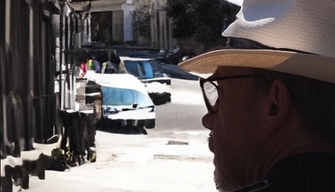 Man in cowboy hat and sunglasses looking down a sunlit alley lined with buildings and parked vehicles.