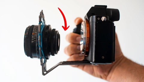 Camera lens mounted on a mirrorless camera body held in a hand, with a red arrow indicating the lens attachment point.