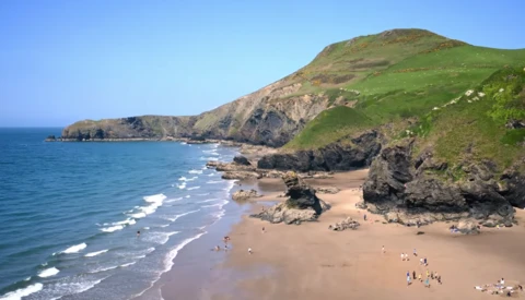 Sandy beach backed by steep green cliffs with rocky outcrops and ocean waves rolling toward shore.