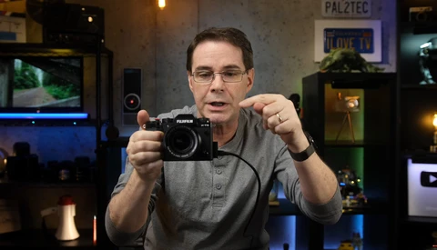 Man in glasses holding a Fujifilm mirrorless camera while speaking to the camera.