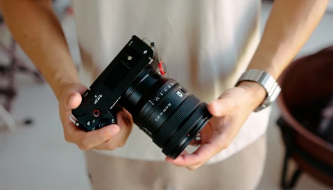Person holding a black rangefinder camera with attached zoom lens against a light-colored shirt.
