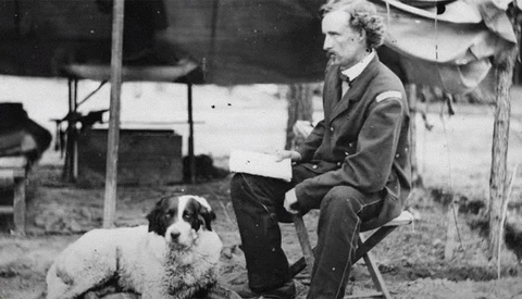 Man in suit seated outdoors holding a document, with a seated dog beside him.