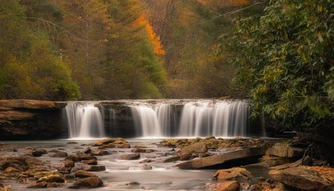 Cascading waterfall flowing over rocky ledges with autumn foliage in background.