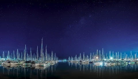 Sailboat masts and rigging reflected in still harbor water at night under a starry sky.