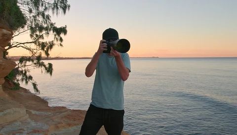 Photographer holding binoculars to eyes while standing on a beach at sunset.