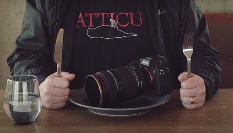 Photographer holding utensils and camera in dining setup, wearing ATTICU branded shirt.