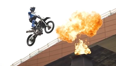 Motocross rider airborne on dirt bike beside massive explosion of flames.