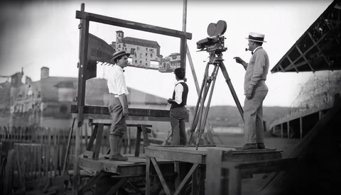 Film crew operating a movie camera on a platform overlooking a European city during early cinema production.