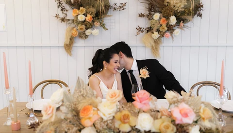 Bride and groom smiling at sweetheart table with floral centerpiece and dried flower wall installation.
