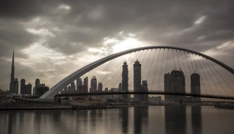 Modern arched bridge spanning a river with city skyline and dramatic cloudy sky reflected in still water.
