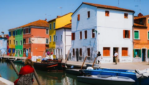 Brightly colored buildings reflected in a canal with moored boats in the foreground.