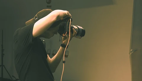 Photographer holding a DSLR camera up to their face, composing a shot in warm studio lighting.