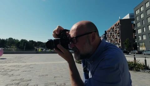 Bald photographer in blue shirt and sunglasses taking a picture with a DSLR camera on a waterfront plaza.